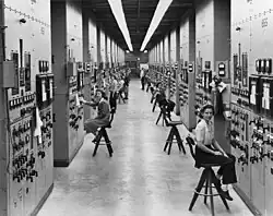 A long corridor with many consoles with dials and switches, attended by women seated on high stools