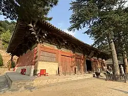 A Chinese temple building. It is built on a raised terrace and two trees nearly obscure the building from view.