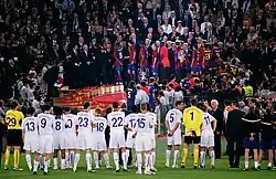 A line of men in white shirts watching men in blue-and-red shirts standing on a rostrum in the background.