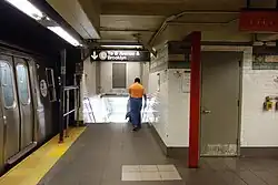 The stairway down to the L train (BMT Canarsie Line) platform at the far south end of the Downtown platform of the 14th Street-Union Square station of the BMT Broadway Line. There is a sign above the staircase, a train at the platform to the left, and a white wall to the right.