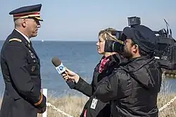 A man in a military uniform is being interviewed by a woman holding a microphone and a man holding a television camera.