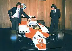 Two men in suits standing left and right of a red-and-white racing car