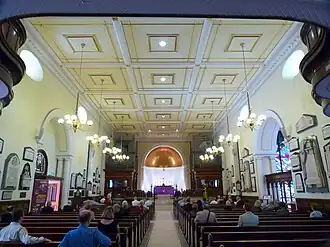 A symmetrical photo looking down the nave towards the apse and chancel showing the corners of the underside of the western gallery at its edges and the pews with some people sitting in them