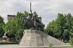 A massive statue of a man riding a horse, set atop a heavy gray stone pedestal against a city background.