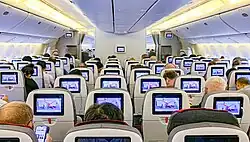 Light gray airliner cabin. Rows of seats arranged between two aisles. Each seatback has a monitor; light shines from the sidewalls and overhead bins