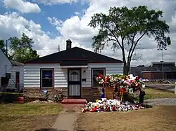 Front of the small white wooden bungalow. The lower part consists of bricks. There's a tree next to the house (right side). A narrow path leads to the front door that is flanked by one window on each side. There's a red step in front of the door. On the right lawn you can see a lot of flowers and fluffy toys. The house is cordoned off with a yellow line.