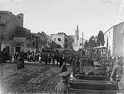 Towers and buildings some with people looking down from roofs with local people and soldiers in a large square with motor car in foreground