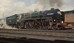 A steam locomotive pictured releasing lots of steam from quite low on the ground, with some unoccupied tracks in between. It is a colour photo