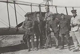 Full-length outdoor portrait of six men in military uniforms in front of a military biplane with a machine gun mounted on the upper wing
