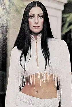 Cher stands in a white, fringed crop top with long sleeves and a white skirt, her long black hair parted in the middle and her midriff exposed.