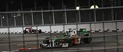 a white, green, and orange F1 car drives down along a closed street at night ahead of a silver F1 car