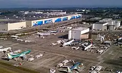 An aerial photograph of a large factory and an airport with several jetliners surrounded by equipment and ground vehicles.
