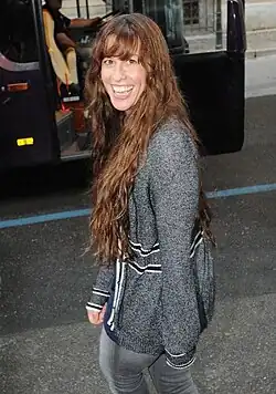 Alanis Morissette smiles while walking on a street. She is looking back over her left shoulder. She wears a sweater and jeans. In the background, there is a bus.