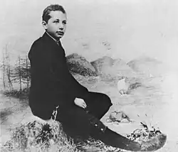 Studio photo of a boy seated in a relaxed posture and wearing a suit, posed in front of a backdrop of scenery.