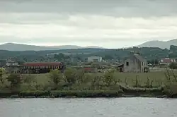 A collection of ruined farm buildings and a nissen hut sit amid fields that lie beyond the grey waters of a river. Wooded slopes and higher treeless hills lie beyond.