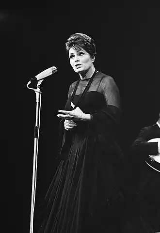 Black-and-white photograph of Amália Rodrigues singing into a standing microphone, wearing a long dark gown and shawl, with one hand raised in expression and a guitarist partly visible in the background.