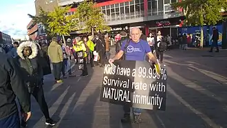 A man in public holding a sign reading "Kids have a 99.998% Survival rate with NATURAL immunity".