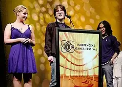 Two men stand behind a podium marked Independent Games Festival, while a woman in a blue dress stands to the left.