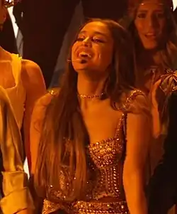 Grande wearing a metallic gold embellished top and skirt with her hair down, smiling under warm yellow lighting