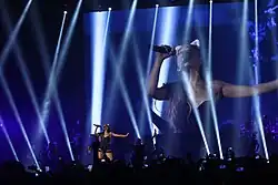 Photograph of Grande performing on stage during the Honeymoon Tour, wearing a black outfit and cat ears, illuminated by multiple white spotlights with a large projection of her face on the screen behind her