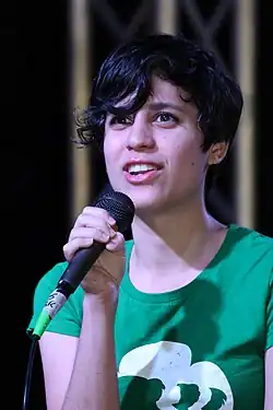 A 24-year-old woman with dark hair and a green shirt looking to the left of the camera and holding a microphone