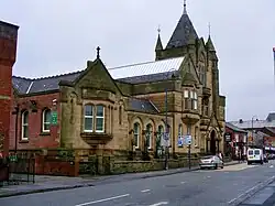 The façade of Ashton town library, constructed from stone and built in Gothic revival style. The photo is a colour modern photo taken from across the road.