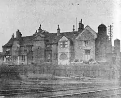 A smudged black-and-white photo of a manor house sitting behind some railway tracks, in front of which the photo was taken. The manor appears medieval in style but has a bric-a-brac façade of various extensions and components.