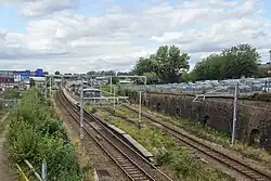 An electrified railway station which has two tracks with an island platform in the middle. The station is quite overgrown both on the platform and on the other sides of the tracks, and there is a steep cutting wall on the right-hand side