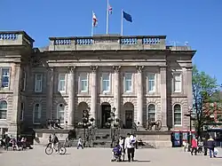 A panoramic shot of a town. In the foreground a modern-looking bus station in front of a red-brick building with large sash windows; in the background a church spire and a hill with houses on the hillside.