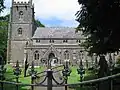 Gray stone building with square tower at left hand end. Foreground includes grass area with gravestones, taken over the top of metal railings.