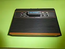 Closeup of a black flat electronic device with a black plastic video game cartridge inserted into the receptacle slot on top. The slot is flanked on both sides by sets of two switches. The cartridge's label reads "Space Invaders".
