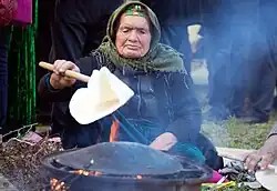 An elderly woman places thin dough onto a convex griddle over a flame.