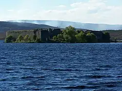 In the foreground are the blue waters of a lake. Beyond that is a wooded shoreline on which there are the ruins of a large walled structure. Smoke drifts across the moorland in the distance.