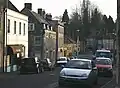 Street scene showing brown stone houses with tiled roofs. Several parked cars in the road.