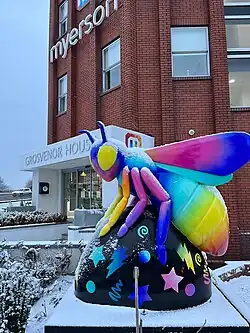 A brightly coloured bee sculpture covered lightly in snow, positioned outside the entrance of Grosvenor House, home to Myerson Solicitors in Altrincham. The sculpture features rainbow wings and limbs with a black base decorated with stars, lightning bolts and other vibrant patterns. Snow blankets the surrounding bushes and ground, with the red-brick office building in the background.