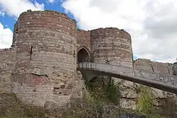 Two round towers of light yellow stone at the bottom and dark orangy stone at the top on either side of an arched entrance. A bridge leads from the entrance to allow access.