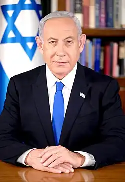 Portrait of Benjamin Netanyahu, a middle-aged man, wearing a suit with a blue tie, sitting in front of an Israeli flag