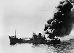 A ship burning from the stern settles in the water.