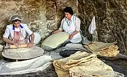 A woman rolls out dough while another stretches dough over a cushion, sitting next to an oven in the ground. Next to her is a stack of finished lavash.