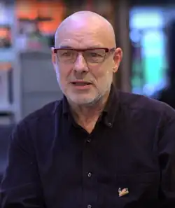 An old man wearing glasses and a dark blue shirt is interviewed