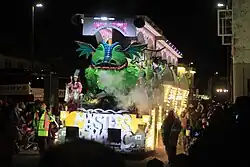 Night time photograph of lorry and trailer decorated with hundreds of coloured light bulbs passing through a street lined by crowds of people.
