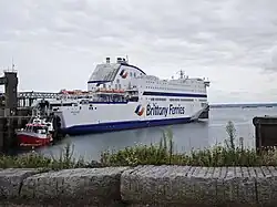 A large Brittany Ferries vessel, the Armorique, docked at a quiet harbor under an overcast sky, with a smaller red service boat positioned near its bow.