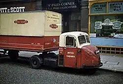An old-fashioned small articulated truck photographed from the side. It is sitting in front of a shop parked parallel close to the curb
