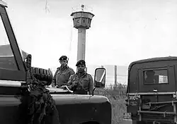 Two British soldiers carrying rifles standing behind a pair of Land-Rover vehicles, one of which has a "British Frontier Service" plate. Behind them is a high mesh fence, behind which is a tall watchtower with an octagonal cabin at its top.