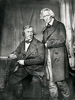 Daguerreotype photograph showing Wilhelm Grimm seated at a table holding a book, with Jacob Grimm standing beside him, taken in 1847.