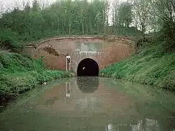 Red brick tunnel entrance through which light can just be seen at the far end. On either side are grassy banks down to the water.