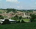 multiple buildings including a square church tower amongst fields and trees.