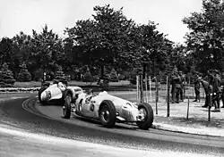 A front engined single-seated racing car leads a similar rear-engined car in a race.