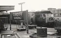 A black-and-white photograph of a railway platform. Two trains stand side by side on the image's right while a sign on the platform reads "Cardiff (General)". A formally dressed gentleman stands on the platform, looking away from the viewer. A briefcase rests on a small trolley beside him