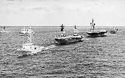 Warships sailing in a line on the open ocean. A small warship leads the line, followed by two aircraft carriers and a replenishment vessel. Other warships sailing in the same direction are in the background.